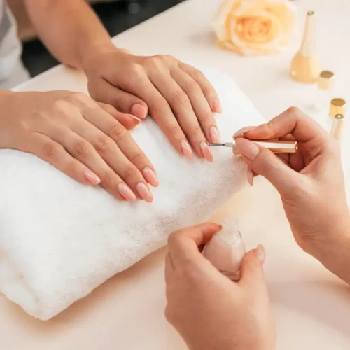 Manicura en proceso con esmalte nude, manos sobre una toalla blanca y productos de belleza visibles en el fondo.