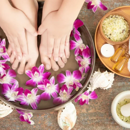 Manos sumergidas en un tazón con agua y pétalos de orquídea, rodeadas de productos de spa como sales de baño, toallas blancas, velas y aceites esenciales, creando un ambiente de relajación y bienestar.